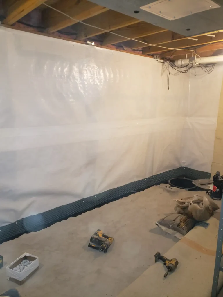 A basement wall with a white plastic barrier suggests a crawlspace encapsulation service in progress; tools and materials are scattered on the concrete floor.