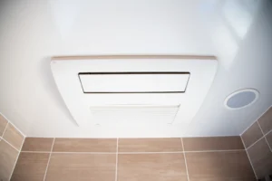 Ceiling-mounted ventilation fan beside a round ceiling light, above beige tiled walls, with seamless dehumidifier installation for optimal moisture control.