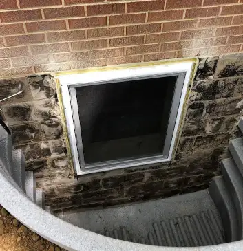 A basement egress window surrounded by stone and concrete is set in a brick wall. Steps lead down to the well-maintained window well. The area appears to be well-lit and clean, suggesting recent repair work for optimal safety and efficiency.