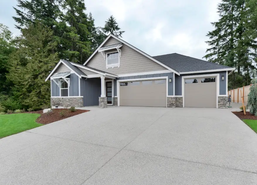 This modern single-story house boasts gray siding and stone accents, with a large driveway and spacious three-car garage. Surrounded by trees, it exemplifies elegance and durability, ensuring lasting beauty even in areas where concrete lifting might be necessary.