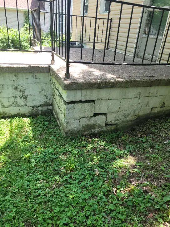 A concrete patio with metal railing displays visible cracks in the foundation blocks, hinting at a need for foundation repair. Lush grass and plants thrive in the surrounding area, adding a touch of nature to the scene.